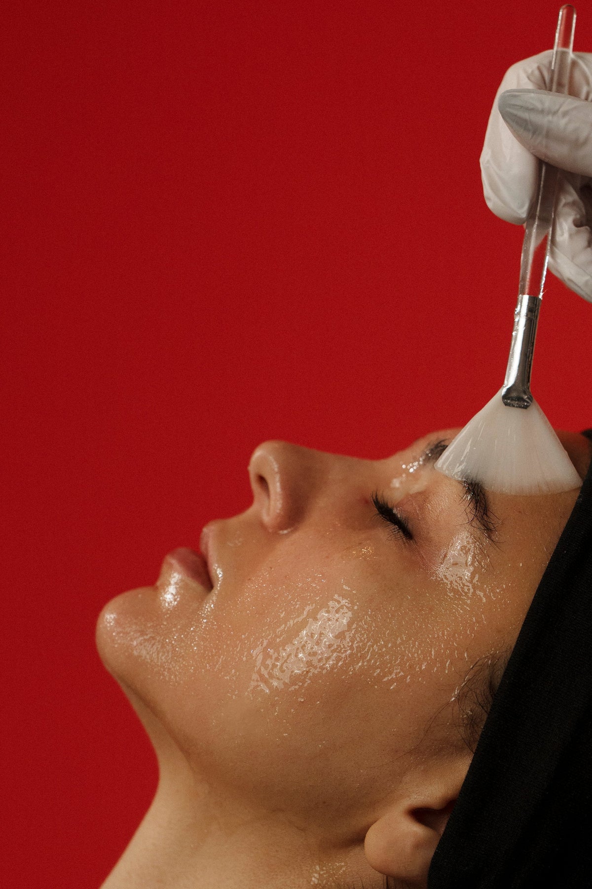 A close-up of a person lying down with their face covered in a glossy white mask or cream, while a gloved hand holds a spatula above their forehead. The background is a solid bright red.